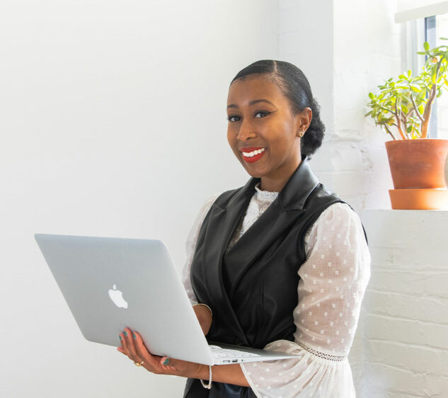 A smiling woman holds a MacBook in front of a small indoor plant