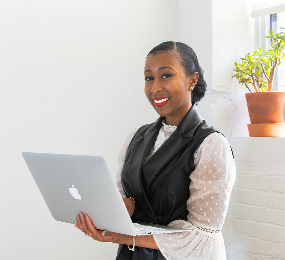 A smiling woman holds a MacBook in front of a small indoor plant